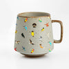 A light gray ceramic mug by Artsy Ems Designs, featuring a brown rim and handle, is adorned with vibrant bird illustrations scattered across its surface—an artistic charm for nature lovers, shown against a plain white background.