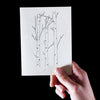 A hand holds a white Canella cardstock card by Artsy Ems Designs, featuring a minimalist black ink tree trunk and branch illustration, set against a solid black background.