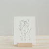 A minimalist black ink drawing of slender tree branches on white Canella cardstock by Artsy Ems Designs, shown upright on a wooden stand against a simple light background.