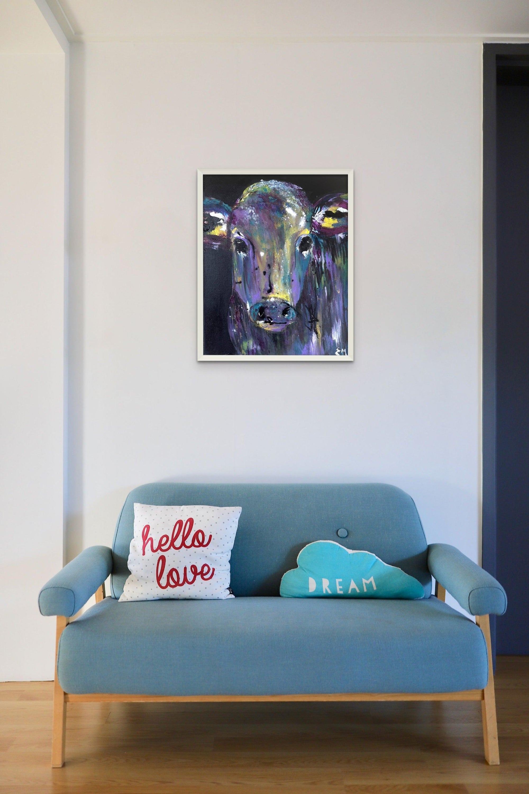 A light blue sofa with hello, love, and dream pillows sits below a colorful cow painting by Artsy Ems Designs—prints available for this vibrant artwork.