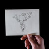 A hand displays a deer drawing with flowers blooming from its antlers, elegantly printed on Canella cardstock by Artsy Ems Designs, set against a plain black background.