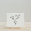 A minimalistic deer head with floral antlers, letterpress printed on crisp Canella cardstock by Artsy Ems Designs. Displayed on a wooden stand against a plain backdrop. Artisan Made in USA.