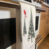 A white 100% cotton kitchen towel by Artsy Ems Designs, featuring pine trees and a red cardinal Christmas motif, hangs on an oven handle with Happy Holidays scripted at the bottom.
