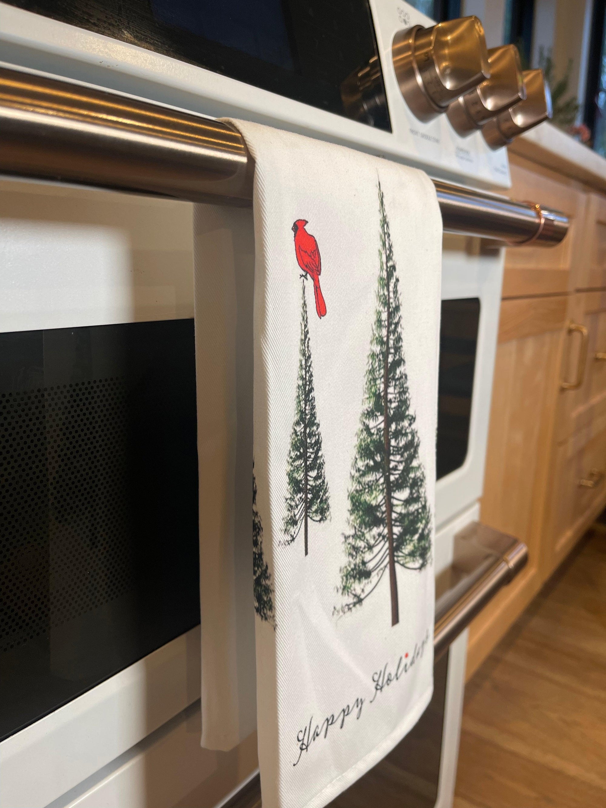 A white 100% cotton kitchen towel by Artsy Ems Designs, featuring pine trees and a red cardinal Christmas motif, hangs on an oven handle with Happy Holidays scripted at the bottom.