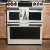A white double oven by Artsy Ems Designs displays a festive “Happy Holidays” towel with pine trees and a red bird on its handle, set in light wood cabinetry and wooden flooring for added seasonal charm.