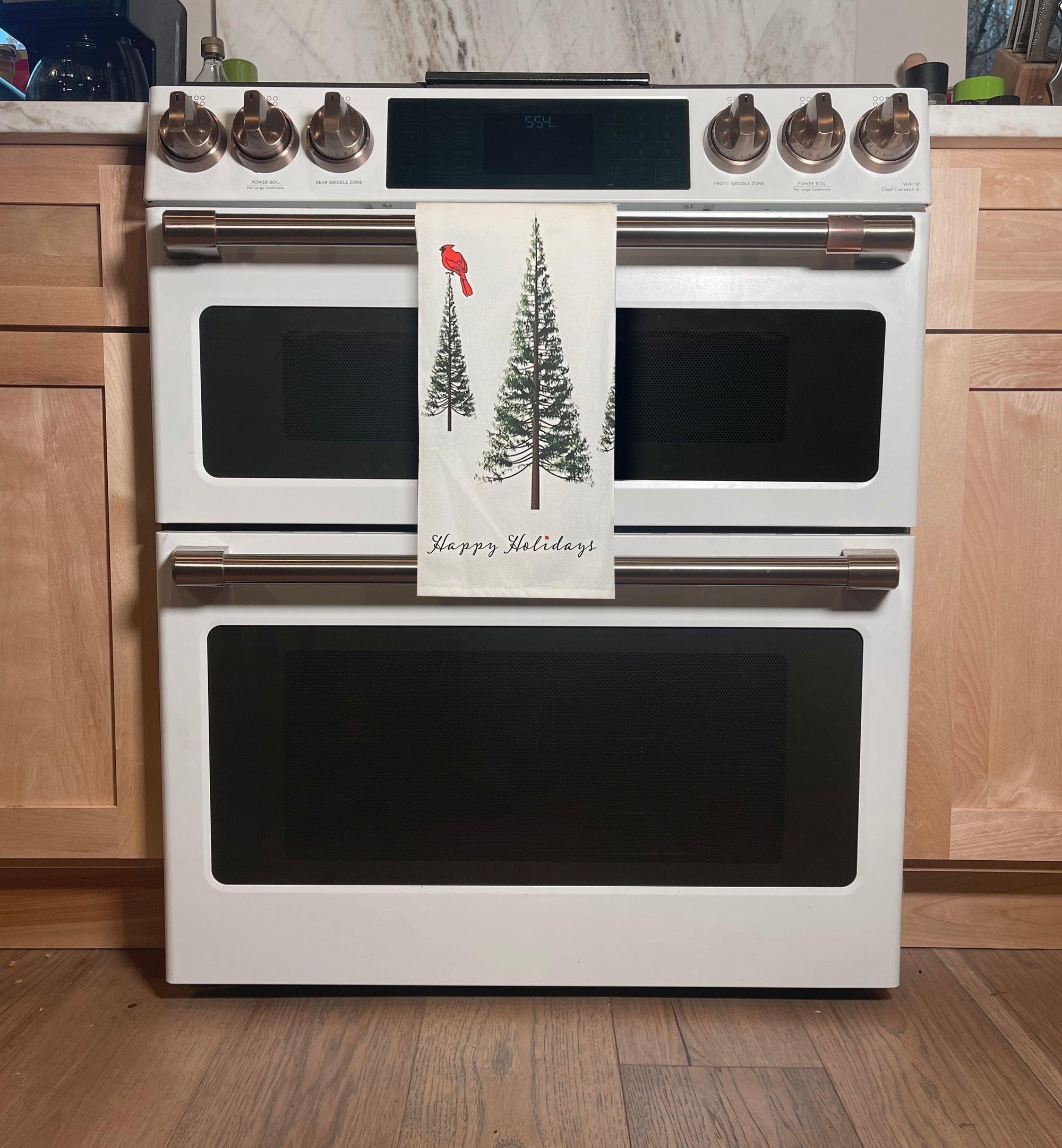 A white double oven by Artsy Ems Designs displays a festive “Happy Holidays” towel with pine trees and a red bird on its handle, set in light wood cabinetry and wooden flooring for added seasonal charm.