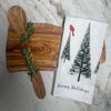 A wooden serving board with two spreaders, rosemary, and a cherry tomato sits beside an Artsy Ems Designs 100% cotton tea towel featuring a Christmas tree and red bird Happy Holidays motif on a marble surface.