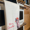 A white 100% cotton kitchen towel by Artsy Ems Designs hangs on an oven handle. It features a Christmas donkey in a Santa hat with red Merry Christmas text. Light wood cabinets and windows are in the background.