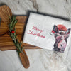 A wooden cutting board with herbs and a red berry sits beside a 100% cotton Artsy Ems Designs white cloth featuring a donkey in a Santa hat and Merry Christmas, displayed on a marble surface.