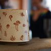 An Artsy Ems Designs Woodland Mug, featuring artisan-crafted brown mushroom and plant illustrations, sits on a counter—its enchanting charm making it the perfect gift for nature lovers. Blurry figures appear in the background.